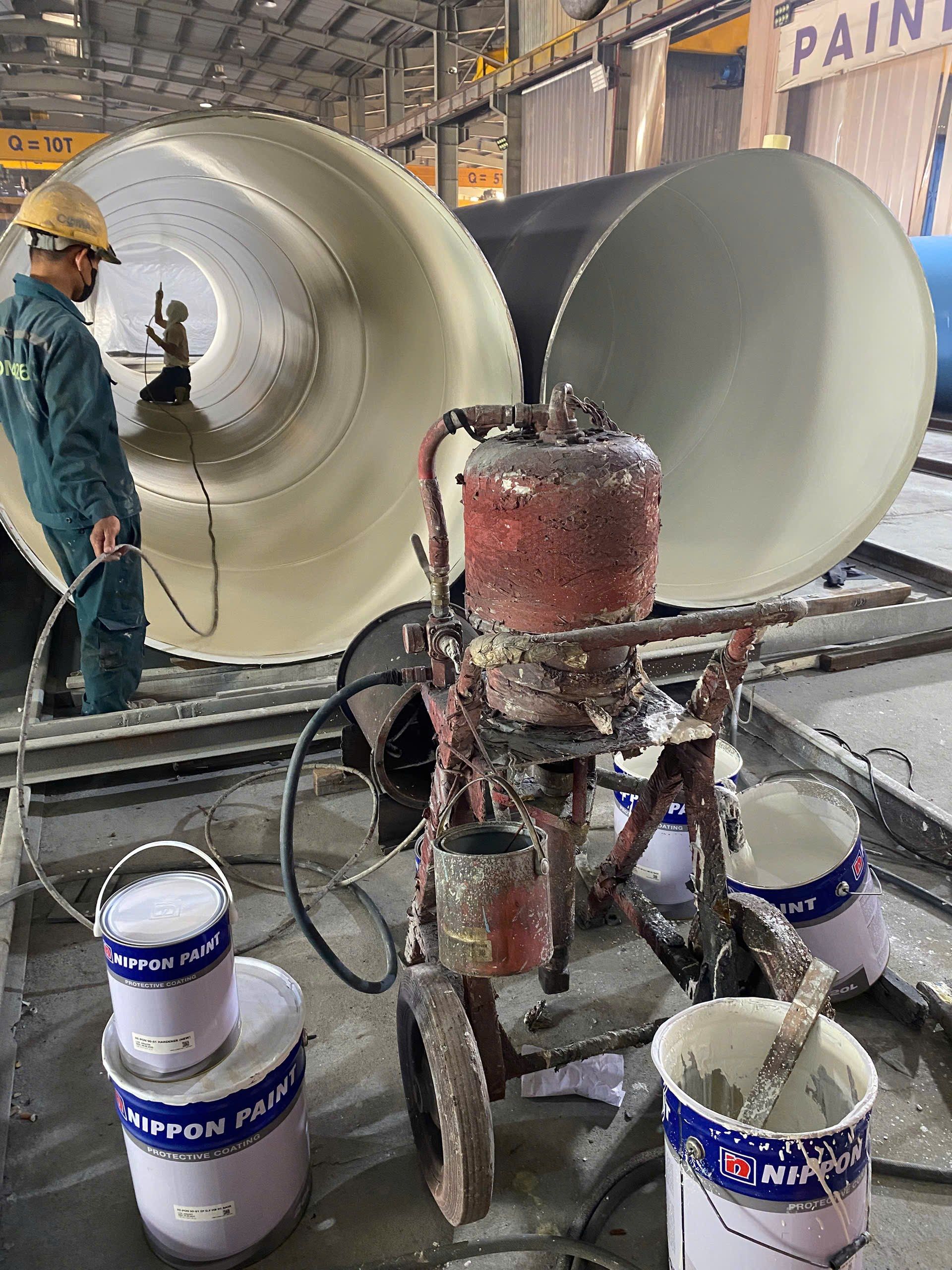 Thợ phun sơn mặt trong ống thép cỡ lớn bên cạnh các thùng sơn